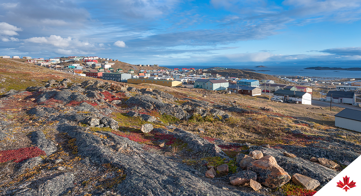 Une vue panoramique d’Iqaluit, capitale du Nunavut, qui comprend un terrain rocheux et une végétation clairsemée figure au premier plan. Des bâtiments colorés parsèment le milieu de l’image, nichés parmi les collines ondulantes sous un ciel partiellement nuageux. Au loin, la baie Frobisher s’étend à l’horizon, évoquant le rôle de la ville comme centre culturel et administratif de l’Arctique.
