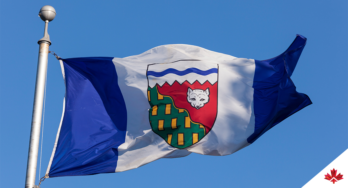 Image du drapeau des Territoires du Nord-Ouest flottant dans un ciel dégagé. Le drapeau affiche deux panneaux latéraux bleu foncé et un large panneau central blanc représentant l’eau, la glace et la neige du Nord. Au centre se trouve l’écu territorial, surmonté d’une section blanche barrée d’une ligne bleue ondulée symbolisant la banquise polaire et le passage du Nord-Ouest. Une ligne diagonale sépare une zone verte avec des rectangles d’or, signifiant les régions boisées et la richesse minérale, et une zone rouge avec un renard arctique blanc, un clin d’œil au patrimoine commercial historique de la région. Ensemble, ces éléments visuels reflètent la géographie et le caractère nordique des Territoires du Nord-Ouest. 