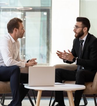 Hommes d’affaires d’ethnies moyen-orientale et caucasienne assis dans un fauteuil dans un bureau moderne en train de discuter.