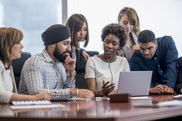 Un groupe diversifié de gens d’affaires se rassemble autour d’un ordinateur portable dans un bureau moderne et discute de ce qu’ils voient.