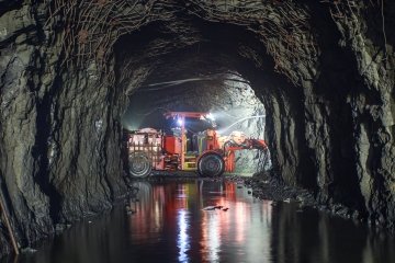 Un véhicule rouge de transport lourd se trouve dans un tunnel souterrain sombre, avec des lumières vives se reflétant sur le sol rocheux et mouillé.