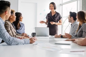 Une femme asiatique d’âge mûr dirige une réunion de collègues de travail multiraciaux discutant ensemble d’idées et de stratégie, assis autour d’une table dans un bureau moderne et créatif en journée, lors d’une réunion du personnel.
