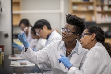Un groupe de techniciens de laboratoire multiethniques travaillant à un bureau, chacun examinant attentivement divers spécimens dans des boîtes de culture cellulaire et discutant des résultats. L’équipement de sécurité et la tenue appropriés sont utilisés.