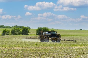 Un tracteur roule dans un champ verdoyant, pulvérisant de l'engrais à partir d'un pulvérisateur monté sous un ciel clair parsemé de nuages. 
