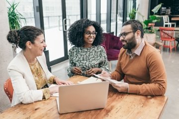 Groupe de professionnels multiraciaux assis à une table, discutant et examinant des plans sur un ordinateur portable. 
