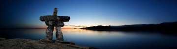 Un grand inukshuk en pierre est bien visible au premier plan, où il se découpe sur un paysage arctique serein au coucher du soleil. Le ciel passe d’un bleu foncé à des teintes chaudes orangées près de l’horizon, qui se reflètent sur les eaux calmes. Les collines lointaines encadrent l’horizon, évoquant l’immensité et la tranquillité du Nunavut.
