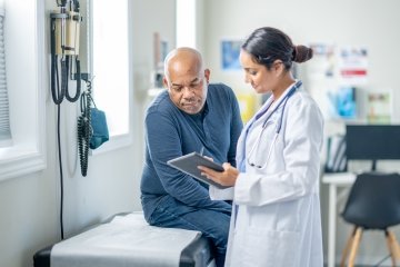 Un homme âgé d’origine africaine est assis sur une table d’examen lors d’un bilan de santé de routine. Sa médecin lui présente une tablette tandis qu’ils examinent ensemble des résultats de tests récents.