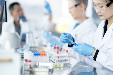 Un technicien de laboratoire médical trie des échantillons de plasma tandis qu’une petite équipe travaille ensemble à les analyser. Ils portent tous des blouses de laboratoire, des lunettes de protection et des gants pendant qu’ils travaillent avec soin.