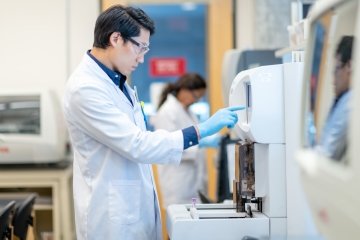Un chimiste asiatique travaille dans un laboratoire. Il se tient devant une grande machine et s’apprête à y insérer un échantillon pour analyse. Il porte des gants de protection et une blouse de laboratoire. Un autre scientifique travaille en arrière‑plan.