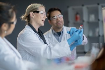 Une petite équipe de personnel de laboratoire médical travaille ensemble sur un vaccin. Ils portent tous des blouses blanches et de l’équipement de protection individuelle tandis qu’ils discutent et étudient divers éléments du vaccin.