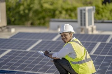 Femme afro-américaine inspectant un toit couvert de panneaux solaires afin d’en assurer l’efficacité, portant un casque et un gilet réfléchissant.